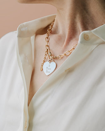 Gold bracelet with heart-shaped tag worn by a person in a light-colored shirt.